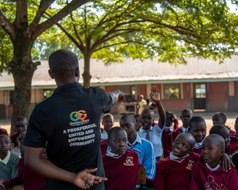 A Gracious Givers Kenya facilitator points to raised hands among schoolchildren during an outdoor session under a large tree.
