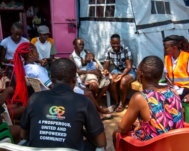 Young people and Gracious Givers Kenya team members in conversation during a community dialogue circle outside market kiosks, one team member wearing a shirt reading “A prosperous, united and empowered community”.