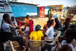 A community dialogue session in a circle of chairs at a market square.