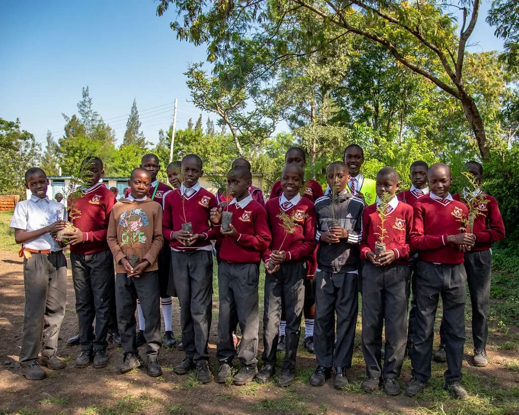 A group of schoolchildren in maroon uniforms stand together under trees, each proudly holding a tree seedling ready for planting.