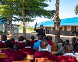 A Gracious Givers Kenya team member waves to seated schoolchildren in a tree-shaded school compound.