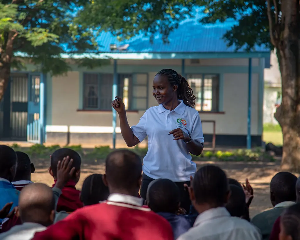 A Gracious Givers Kenya facilitator leads an outdoor session, speaking to schoolchildren seated in front of their school building in Nyando.